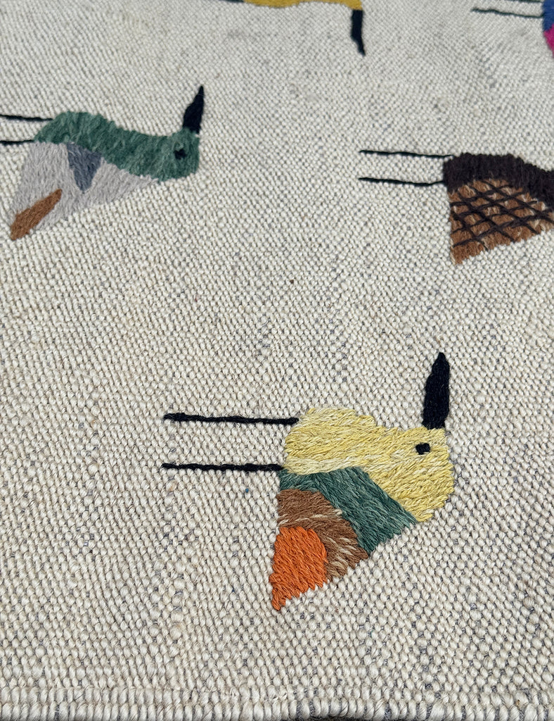 Close-up of a woven suzani rug with a cream base and embroidered bird motifs in muted green, mustard yellow, orange, brown, and black.