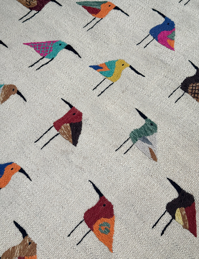 A woven Suzani rug with a light ecru linen-like background, decorated with rows of stylised bird figures in many different colours including blue, red, yellow, orange, green, and black.