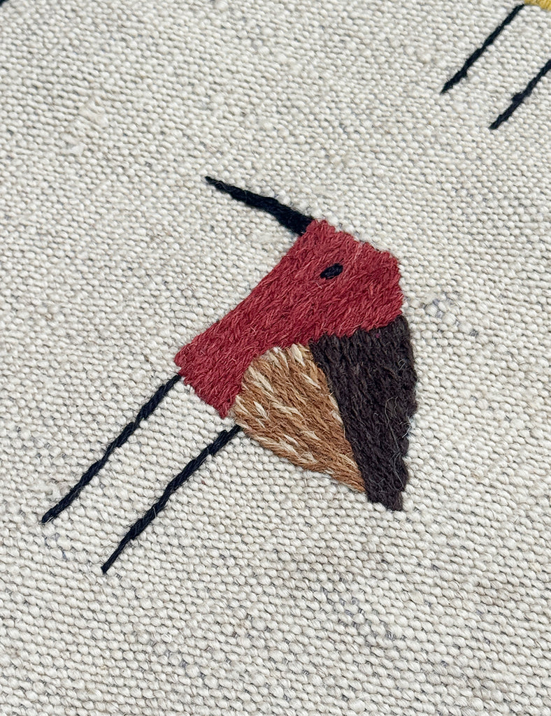 Close-up of a woven textile featuring a stylized embroidered bird motif in red, brown, tan, and black on an off-white textured background.