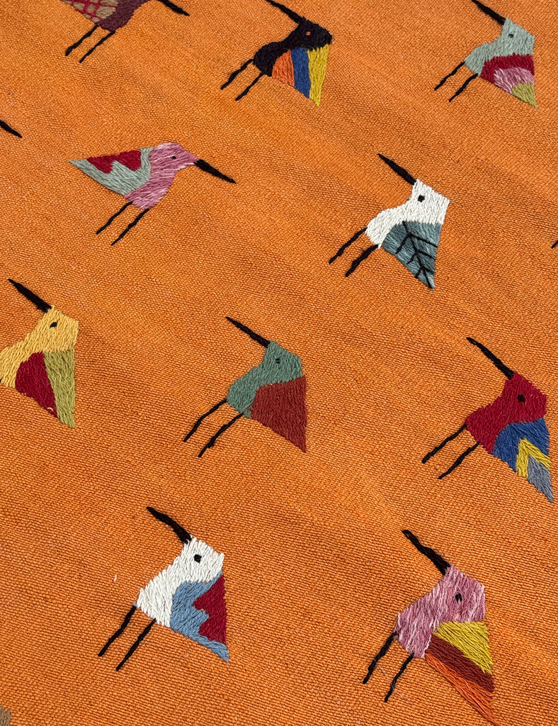 An orange woven SUZANI rug featuring a repeating pattern of stylised birds in various colours including blue, red, yellow, black, and white.