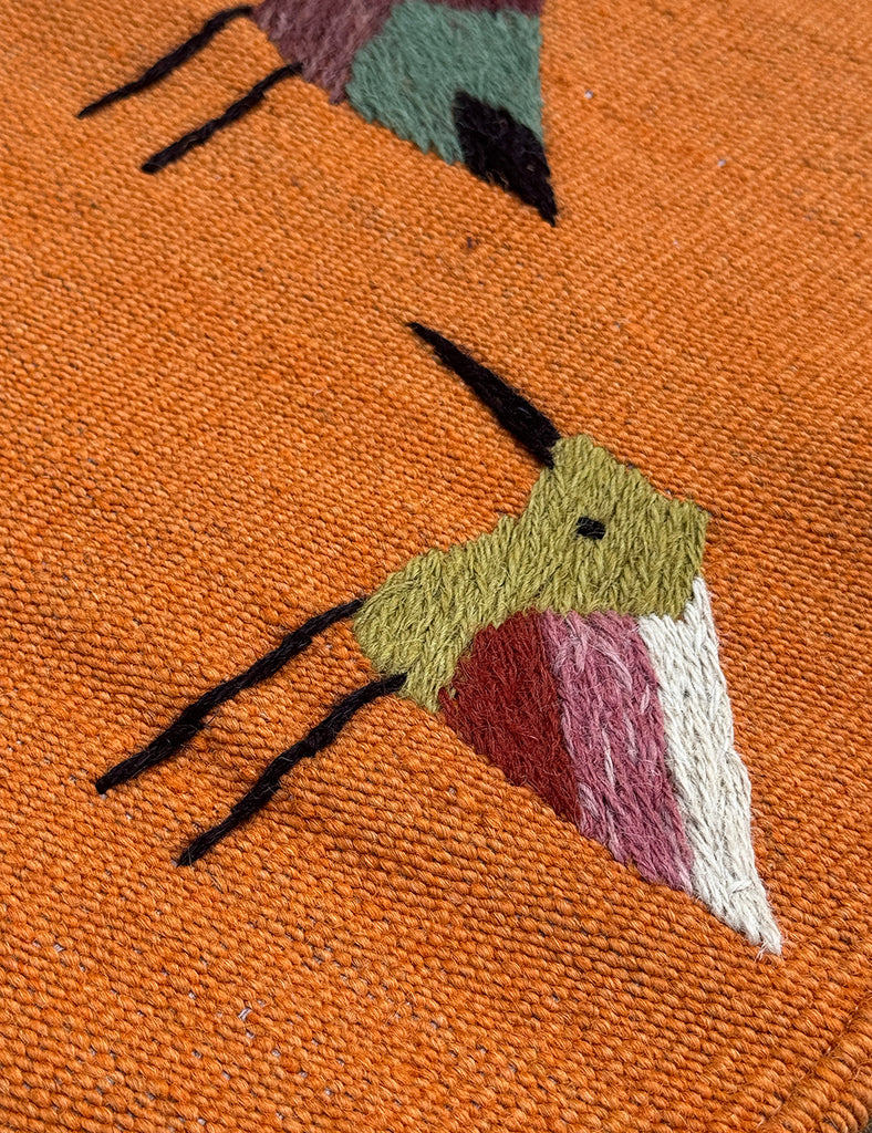 An orange woven SUZANI rug featuring a repeating pattern of stylised birds in various colours including blue, red, yellow, black, and white.